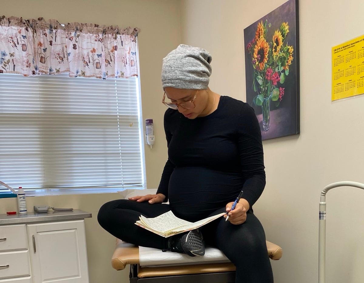 Indira reading from her journal at a doctor's appointment. She is wearing all black with a gray cap and is very pregnant.