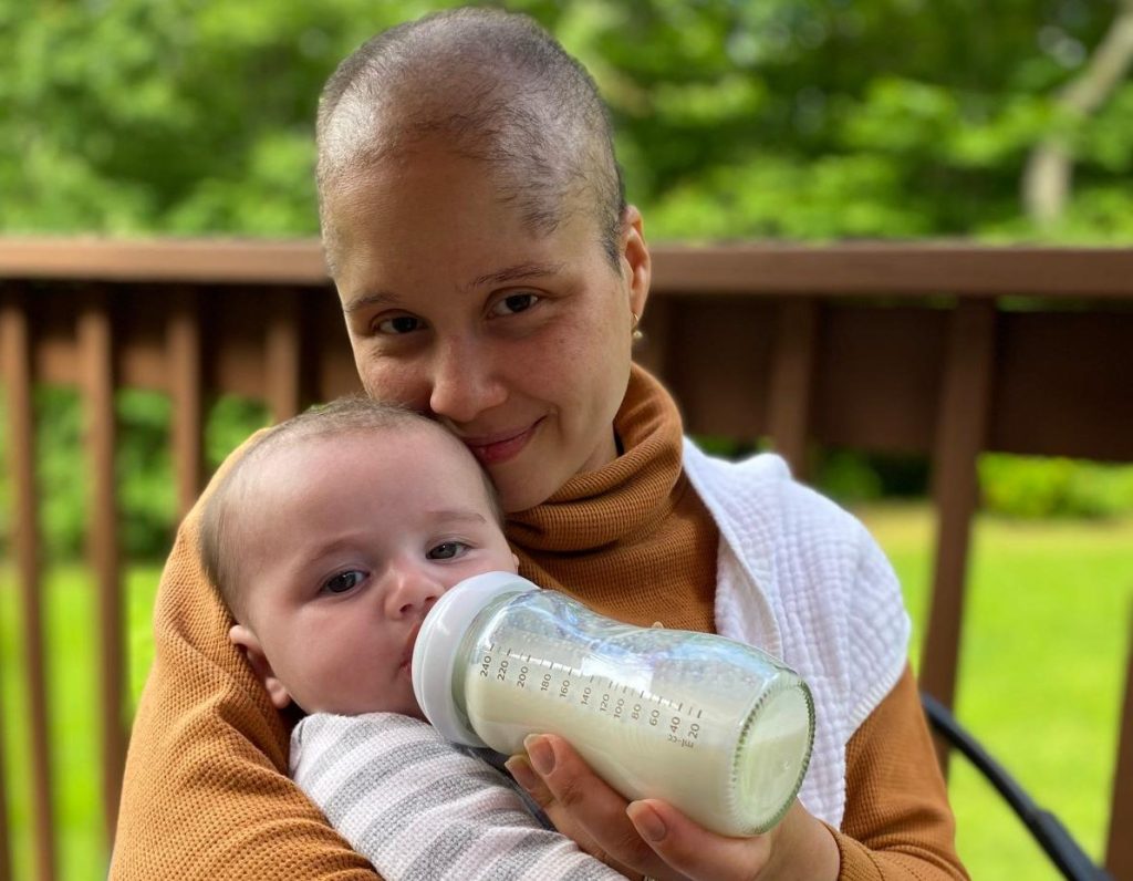 Photo of Indira feeding her five month old baby with a bottle of milk. They are outside. She is smiling and has no hair. The baby is looking at the camera.